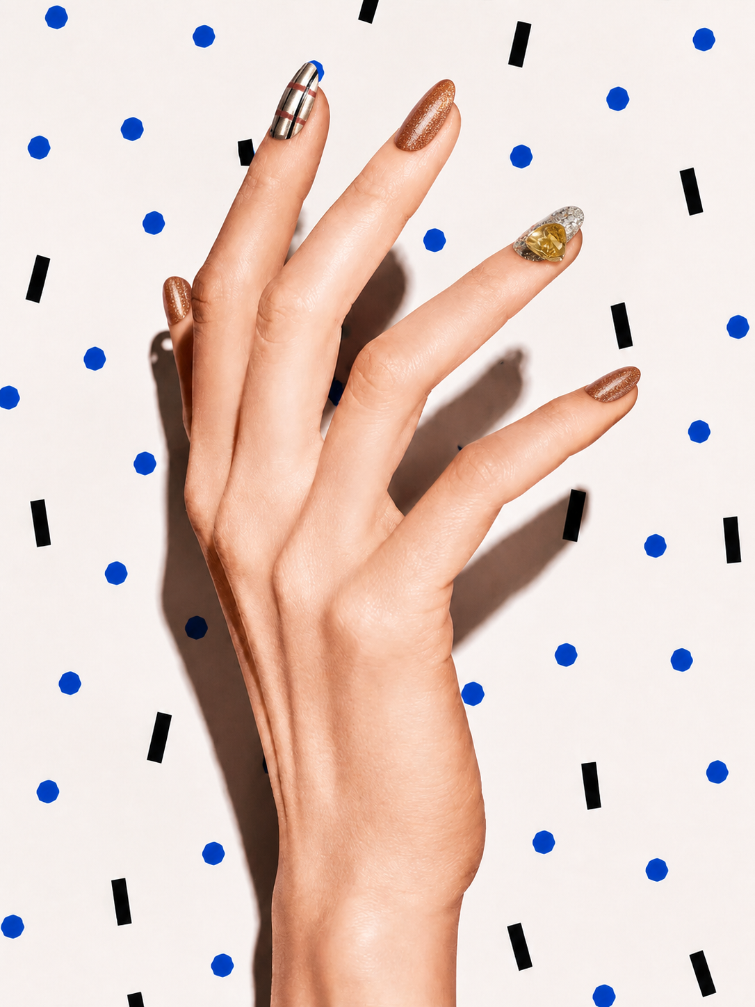 Editorial hand wearing Golden Hour bronze metallic citrine press-on nails on a blue dot patterned background