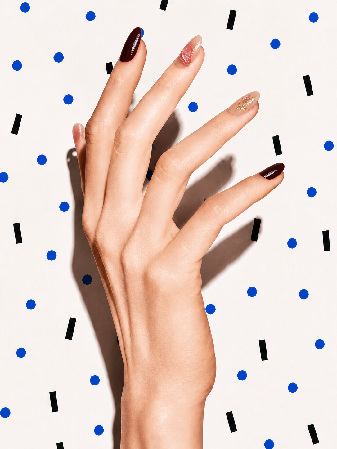 Editorial hand wearing In Bloom burgundy floral rose quartz press-on nails on a blue dot patterned background